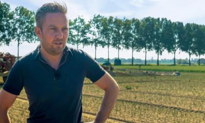 Outdoor agricultural setting with a person wearing a dark polo shirt standing in a field. Trees line the background, and farm equipment is partially visible. The scene is captured in natural daylight with a rural landscape